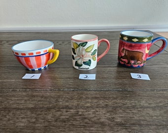 May include: Three ceramic mugs with different designs. The first mug is red and white striped with a yellow handle. The second mug has a white magnolia flower design and the words "Mississippi" at the bottom. The third mug is red with a brown bear design and blue and green accents.