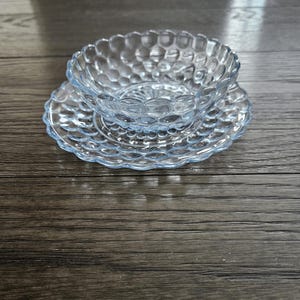 May include: A clear, light blue glass bowl and saucer set. The bowl has a textured, bubble-like pattern, and sits atop a matching saucer with a scalloped edge. The set is displayed on a dark wooden surface.