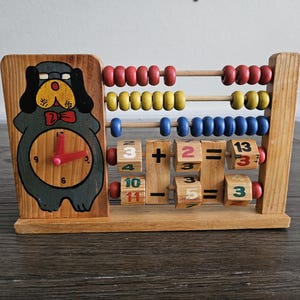 May include: Wooden educational toy featuring a clock and an abacus. The clock has a dog design and red hands. The abacus has red, yellow, and blue beads. Includes wooden blocks with numbers and math symbols.