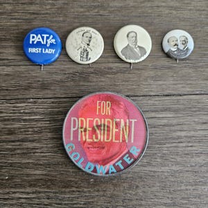 May include: Four vintage political campaign buttons. The top left button is blue with white text that reads "PAT for FIRST LADY". The top middle button is white with a black and white image of a man in a suit and tie. The top right button is white with a black and white image of two men in suits and ties. The bottom button is red with white text that reads "FOR PRESIDENT GOLDWATER" and a red image of a man in a suit and tie.