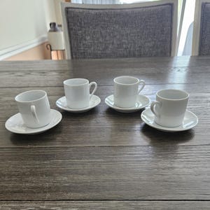 May include: Four white ceramic espresso cups with matching saucers are arranged on a wooden table. Each cup has a handle and sits on a small, round saucer. The cups and saucers are clean and simple in design.
