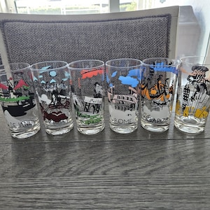 May include: Set of six clear drinking glasses, each featuring a different city scene. Cities include Rio de Janeiro, Paris, Rome, Singapore, and New York. The glasses have colorful illustrations and city names.