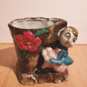 Puede incluir: Una maceta de cerámica con forma de tronco de árbol con una pequeña figura de gnomo sentada en el lateral. El gnomo lleva un atuendo azul y blanco y tiene una flor roja en la cabeza. La maceta está decorada con una flor roja y hojas verdes.