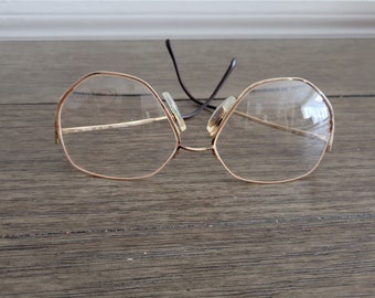 May include: Gold-toned spectacles with a distinctive octagonal frame shape and clear lenses. The glasses are lying on a wooden surface.