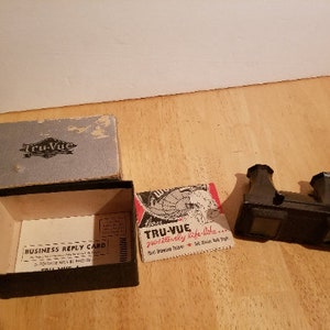 May include: A vintage Tru-Vue 3D viewer with a black plastic body and two viewing lenses. The viewer is accompanied by a box and a business reply card. The card reads "Business Reply Card" and includes an address for Tru-Vue.