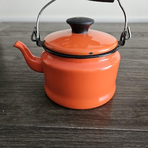 May include: A small, orange enamel teapot with a black handle and lid. The teapot has a silver wire handle and a spout. The design is simple and classic, suitable for kitchen decor.
