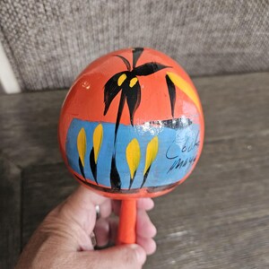 May include: Hand-painted orange maraca with a palm tree design, blue accents, and yellow details. The maraca has a long orange handle and is held in a hand. The text "Coba Maya" is written on the side.