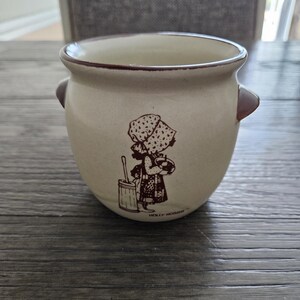 May include: A tan ceramic pot with two small brown handles. The pot features a brown illustration of a girl in a bonnet holding a basket and a bucket. The text "HOLLY HOBBIE" is printed at the bottom of the pot.