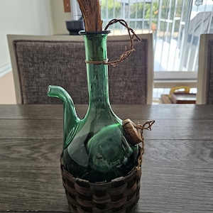 May include: A green glass wine decanter with a woven basket base. The decanter has a spout, a handle, and a natural fiber stopper. The top of the decanter has a bundle of straw. The decanter is sitting on a wooden table.