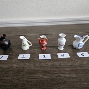 May include: Five miniature ceramic pitchers in various styles and colors. The pitchers are arranged in a row on a wooden surface. The pitchers are numbered 1 through 5.