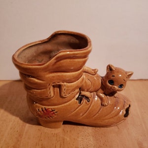 May include: A ceramic boot planter with a brown glaze and a small cat figurine sitting on top. The boot has a red and brown stitched design on the side.
