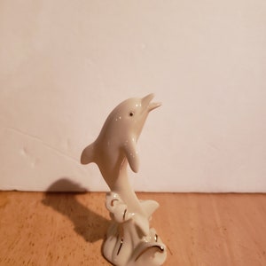 May include: A white porcelain figurine of a dolphin leaping out of the water. The dolphin is painted with gold accents and is set on a base with a wave design.