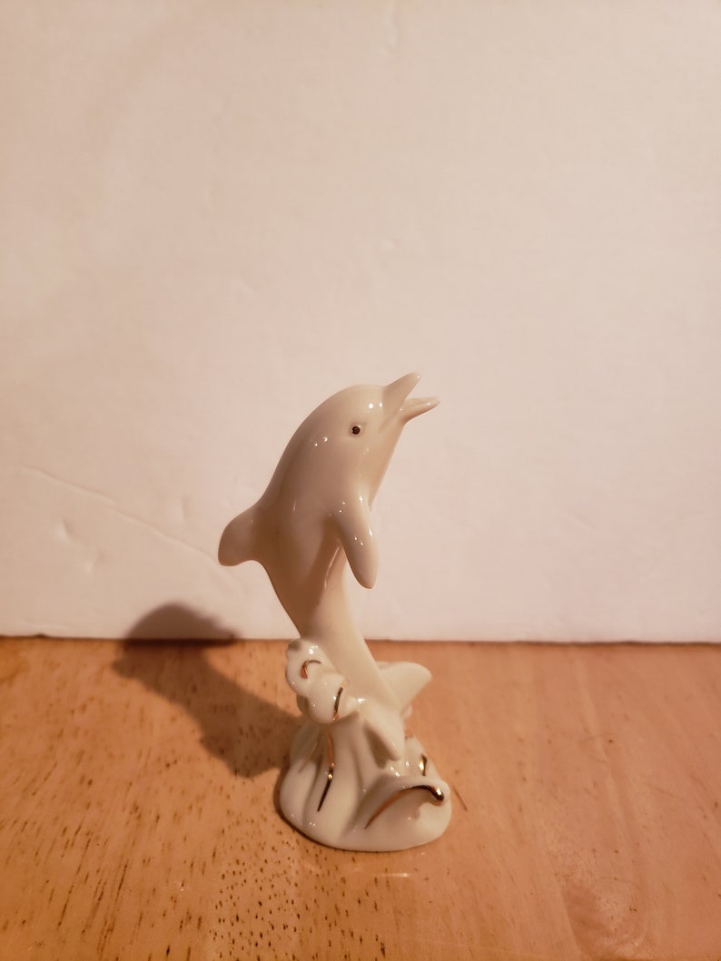 May include: A white porcelain figurine of a dolphin leaping out of the water. The dolphin is painted with gold accents and is set on a base with a wave design.