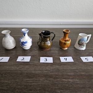May include: Five small ceramic vases in different styles and colors. The vases are arranged in a row on a wooden surface. The vases are numbered 1 through 5.