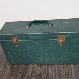 May include: A vintage green toolbox with a handle and brass-colored clasps. The rectangular box has a textured surface and a dark green handle. The clasps and handle hardware show signs of age.