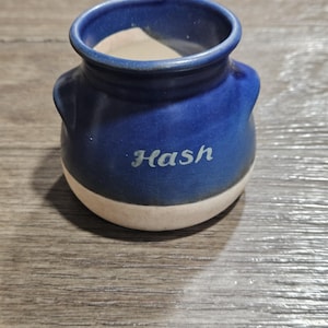 May include: A small, blue ceramic pot with the word "Hash" in white script. The pot has a rounded shape with two small handles and a cream-colored base. The pot sits on a wooden surface.
