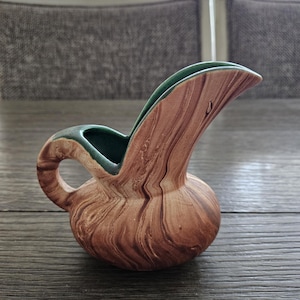 May include: A small, decorative pitcher with a unique shape and a brown and green color scheme. The pitcher has a bulbous base, a curved spout, and a handle. The exterior features a wood-grain pattern, while the interior and rim are green.