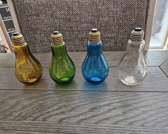 May include: Four decorative glass light bulb-shaped vases in amber, green, blue, and clear. Each vase has a gold-coloured base and a bulb-shaped body. The vases are arranged on a wooden surface.