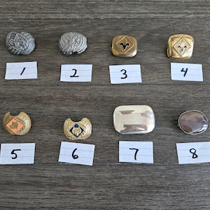 May include: Eight metal and plastic badges with numbers 1 through 8 written below each badge. The badges feature various designs, including a wolf head, a fleur-de-lis, and a ram's head.