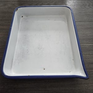 May include: A rectangular white enamel tray with a blue rim. The tray has a slightly tapered shape and is set against a dark wood surface. The tray is empty and shows signs of wear.