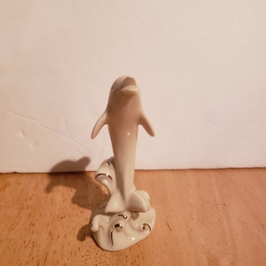 May include: A white porcelain figurine of a dolphin leaping out of the water. The dolphin is detailed with gold accents and is set on a base with a wave design.