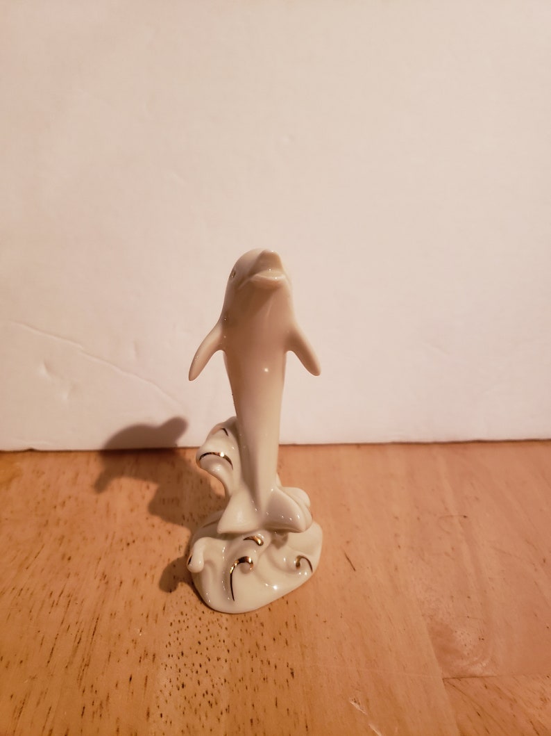 May include: A white porcelain figurine of a dolphin leaping out of the water. The dolphin is detailed with gold accents and is set on a base with a wave design.