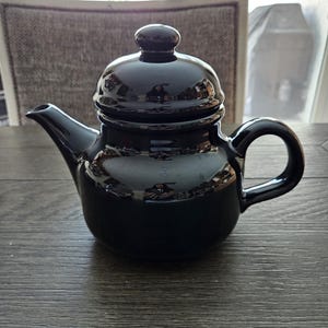 May include: A glossy black ceramic teapot with a matching lid and handle. The teapot has a curved spout and sits on a wooden surface. The teapot is designed for brewing tea.