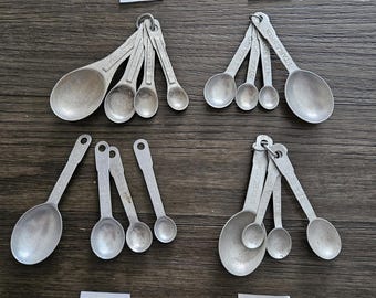 May include: Four sets of vintage metal measuring spoons. Each set includes spoons of varying sizes, connected by a metal ring. The spoons are silver-colored and show signs of wear. The sets are arranged on a dark wooden surface.