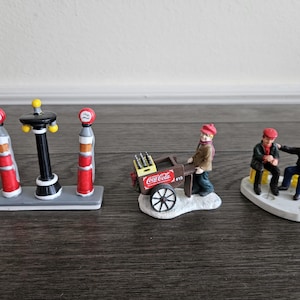 May include: A collection of miniature Christmas village figurines. The set includes a gas station, a Coca-Cola vendor with a cart, and two figures sitting on a bench. The figurines are painted with bright colors and detailed designs.