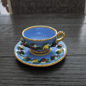 May include: A small, ornate blue teacup and saucer set. The cup and saucer are decorated with a floral pattern in green, red, and yellow. The rim of the cup and saucer is trimmed with gold. The set is placed on a dark wooden surface.
