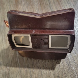 May include: A vintage brown plastic 3D viewer with two rectangular viewing windows. The viewer has a silver trim around the windows and a silver knob on the top.