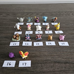May include: A collection of 17 small plastic Pokemon figures, numbered 1 through 17, arranged in rows on a wooden surface. The figures are various colors and depict different Pokemon characters.
