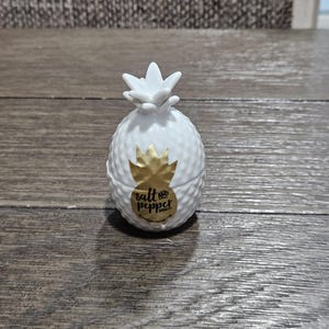 May include: A white ceramic pineapple-shaped salt and pepper shaker set. The shaker has a textured surface and a gold label that reads "salt and pepper". The pineapple top is white and has a star-like shape.