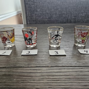 May include: Four vintage shot glasses with gold rims, each featuring a different cartoon character design. The glasses are numbered 1 through 4 on small white labels. The shot glasses are on a wooden surface.