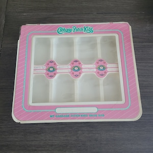 May include: A pink and white plastic storage box with a clear plastic window. The box has a label that says "Cabbage Patch Kids" and "My Cabbage Patch Kids Brag Bag". The window shows six compartments for storing items.