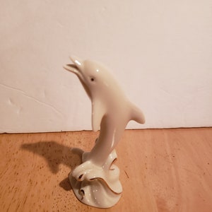 May include: A white porcelain figurine of a dolphin leaping out of the water. The dolphin is painted with gold accents and is standing on a base that resembles a wave.
