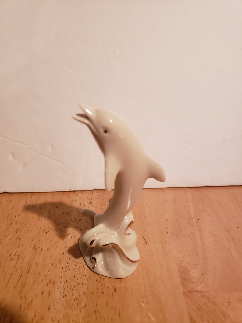 May include: A white porcelain figurine of a dolphin leaping out of the water. The dolphin is painted with gold accents and is standing on a base that resembles a wave.