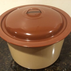 May include: A brown enamel pot with a lid. The pot has a beige exterior and a brown interior. The lid has a brown exterior and a beige interior. The pot has a handle on the lid.