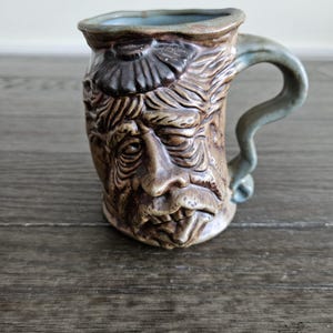 May include: A brown ceramic mug with a sculpted face design. The mug features a detailed depiction of a bearded face with furrowed brows and a textured surface. The handle is a light green color, and the interior rim is light blue.