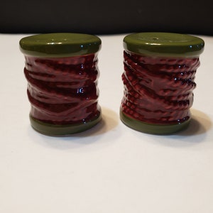 May include: A set of two ceramic salt and pepper shakers shaped like spools of thread. The shakers are red with a green top and bottom.
