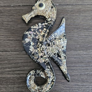 May include: A decorative seahorse figurine with a black body and iridescent accents. The seahorse is embellished with small, irregularly shaped pieces in shades of beige and gold. The eye is a light beige color. The seahorse is approximately 8 inches long.