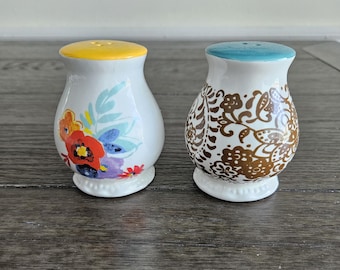 May include: Two white ceramic salt and pepper shakers. One shaker has a yellow top and floral design with red, orange, and purple flowers. The other shaker has a blue top and a brown floral pattern. Both have a white base.