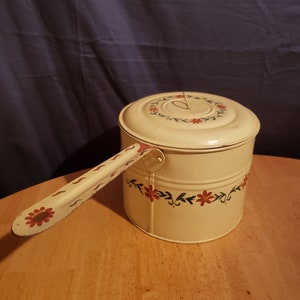 May include: A vintage white enamelware pot with a floral design in red and green. The pot has a lid and a handle.