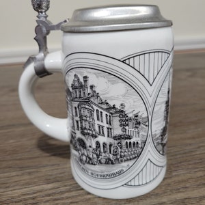 May include: White ceramic beer stein with a pewter lid and a black and white illustration of a building with the text "Muenchen Hofbrauhaus".