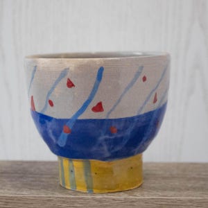 Può includere: Una piccola ciotola in ceramica con un design a strisce blu e bianche e accenti rossi. La ciotola ha una base gialla con strisce blu e bianche.