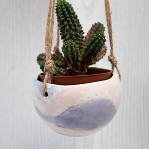 May include: A hanging planter made of white ceramic with a light blue and pink abstract design. The planter is hanging from a natural twine rope and contains a small green succulent plant.