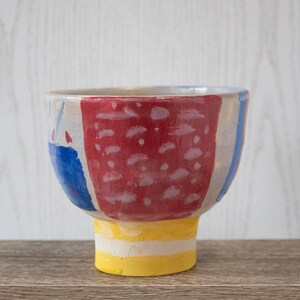 Puede incluir: Un cuenco de cerámica con una base amarilla y un diseño pintado en rojo, azul y blanco. El cuenco tiene una abertura ancha y redonda y una base estrecha.