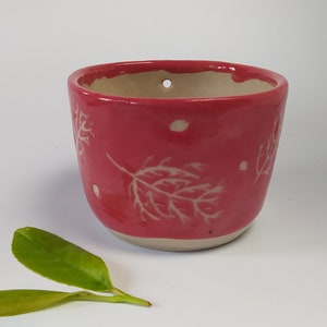 Puede incluir: Un pequeño macetero de cerámica rojo con un diseño de hojas blancas. El macetero tiene un agujero de drenaje y es perfecto para colgar.