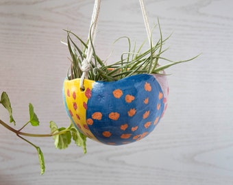 May include: A colorful ceramic hanging planter with a blue, yellow, and pink geometric pattern. The planter is filled with green air plants and is hanging from a white string.