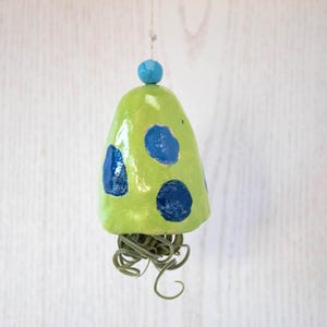 Puede incluir: Una campana de cerámica verde con lunares azules y una cuenta azul en la parte superior. La campana está colgada de una cuerda y tiene una planta de aire verde colgando de la parte inferior.